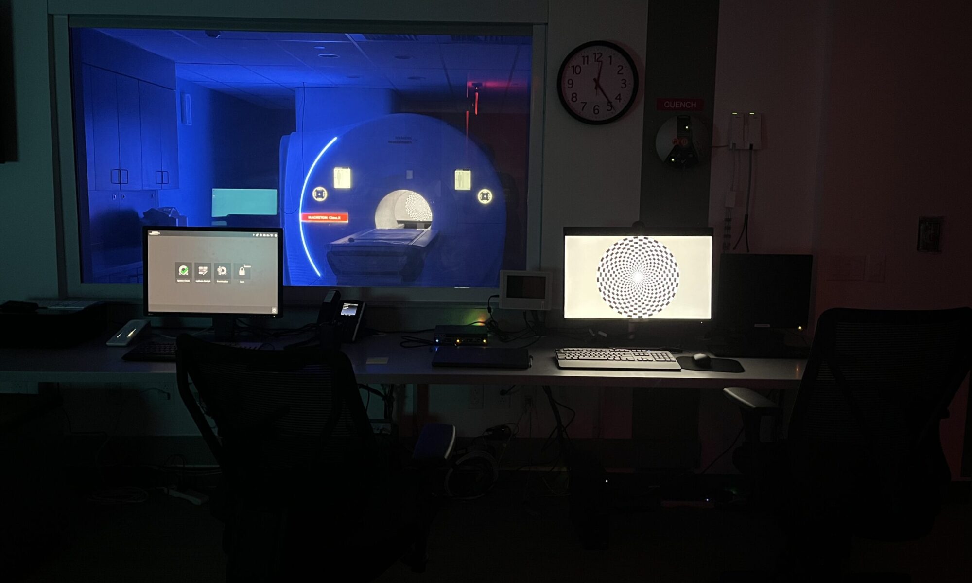 An MRI scanner as seen from the control room. Lighting is dramatic as all the room lights are off and only the scanner mood lighting and control computers are visible.